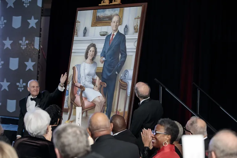 a man in tuxedo gestures to a large portrait of the Wrightons